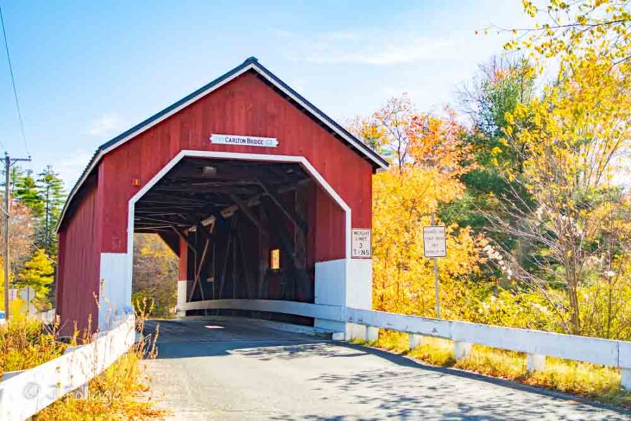 Small red Carlton Covered Bridge spanning a quiet tributary with peak New Hampshire fall foliage.
