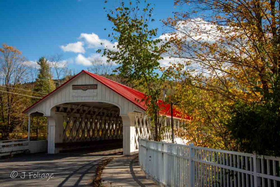 White Ashuelot Covered Bridge with a red roof and open lattice sides reflecting in the river.