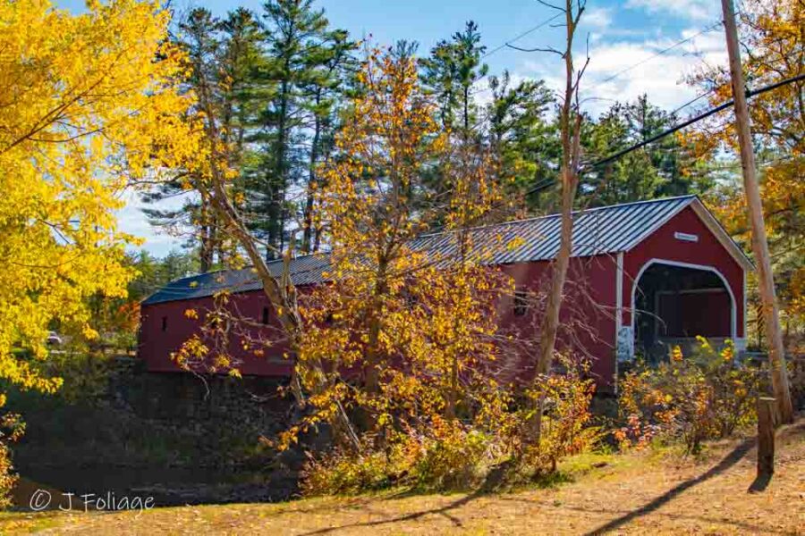 Red Sawyers Crossing Covered Bridge viewed through autumn trees over the Ashuelot River in Swanzey, NH.