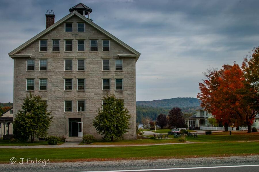The four-story granite Great Stone Dwelling at Enfield Shaker Village in New Hampshire, framed by vibrant orange and red autumn foliage under a soft blue sky.