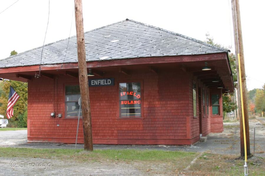 The historic dark wood Old Enfield railroad station building, featuring a red "Enfield" sign, surrounded by lush green trees.