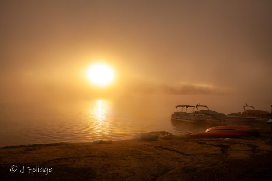 A serene sunrise view of Mascoma Lake in Enfield, New Hampshire, with morning mist rising off the water and golden light reflecting on the surface.
