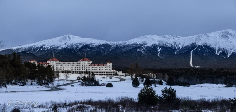 Winter View: The grand white facade and iconic red roof of the Omni Mount Washington Resort sitting at the base of the snow-capped Presidential Range.